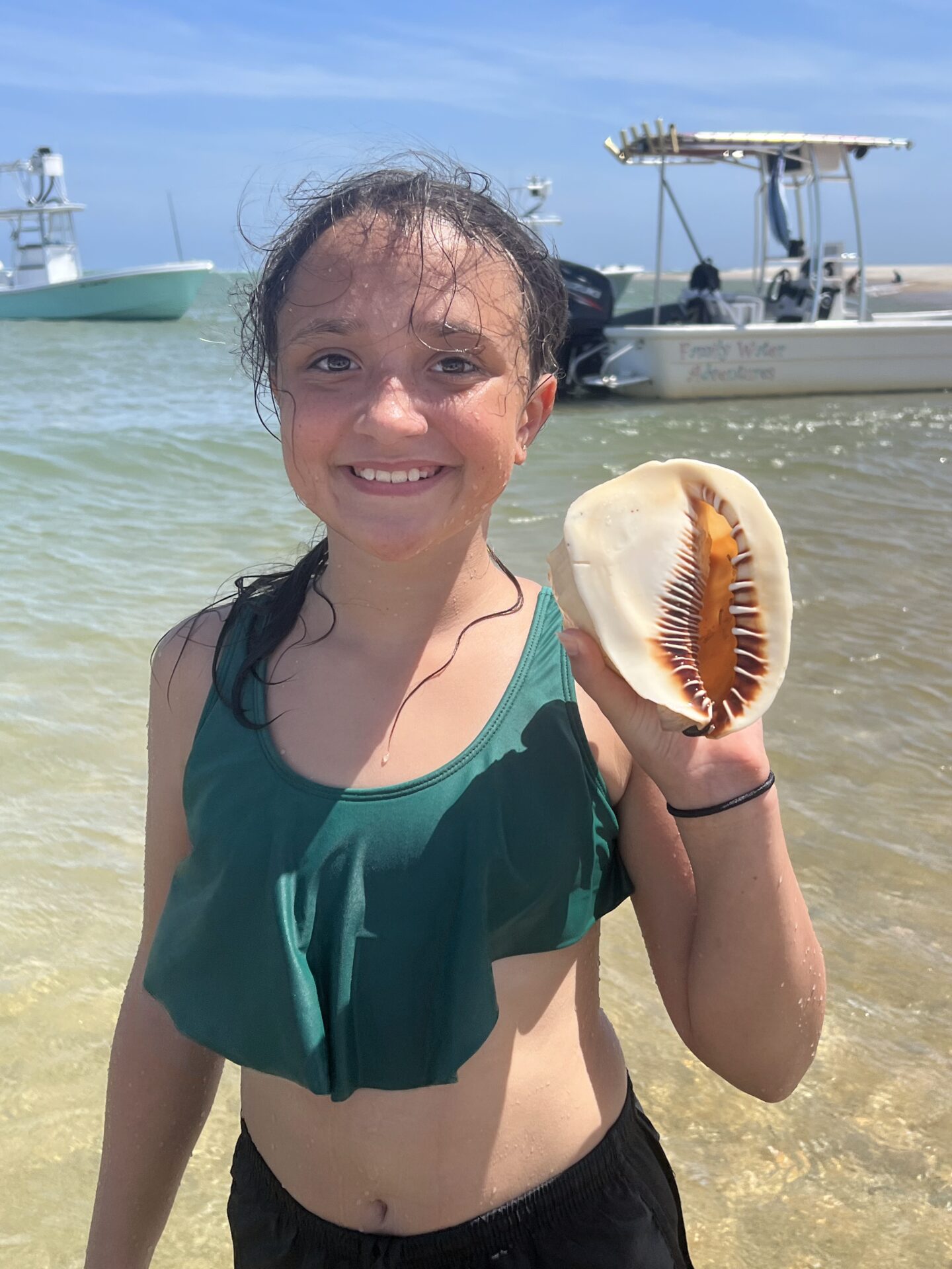 Smiling girl holding a large seashell on the beach with boats in the background.