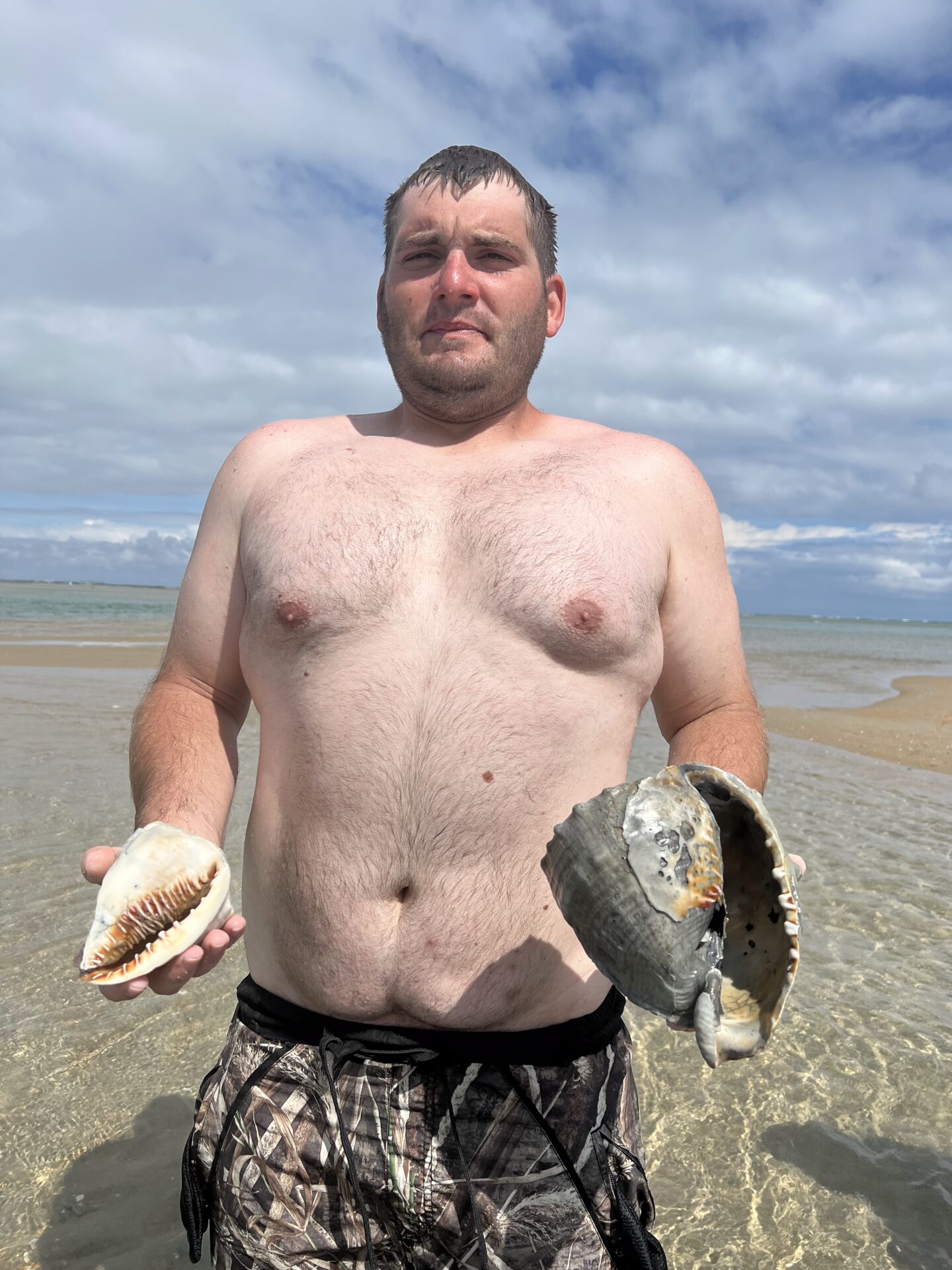 Man holding two large fish on a beach under cloudy sky.